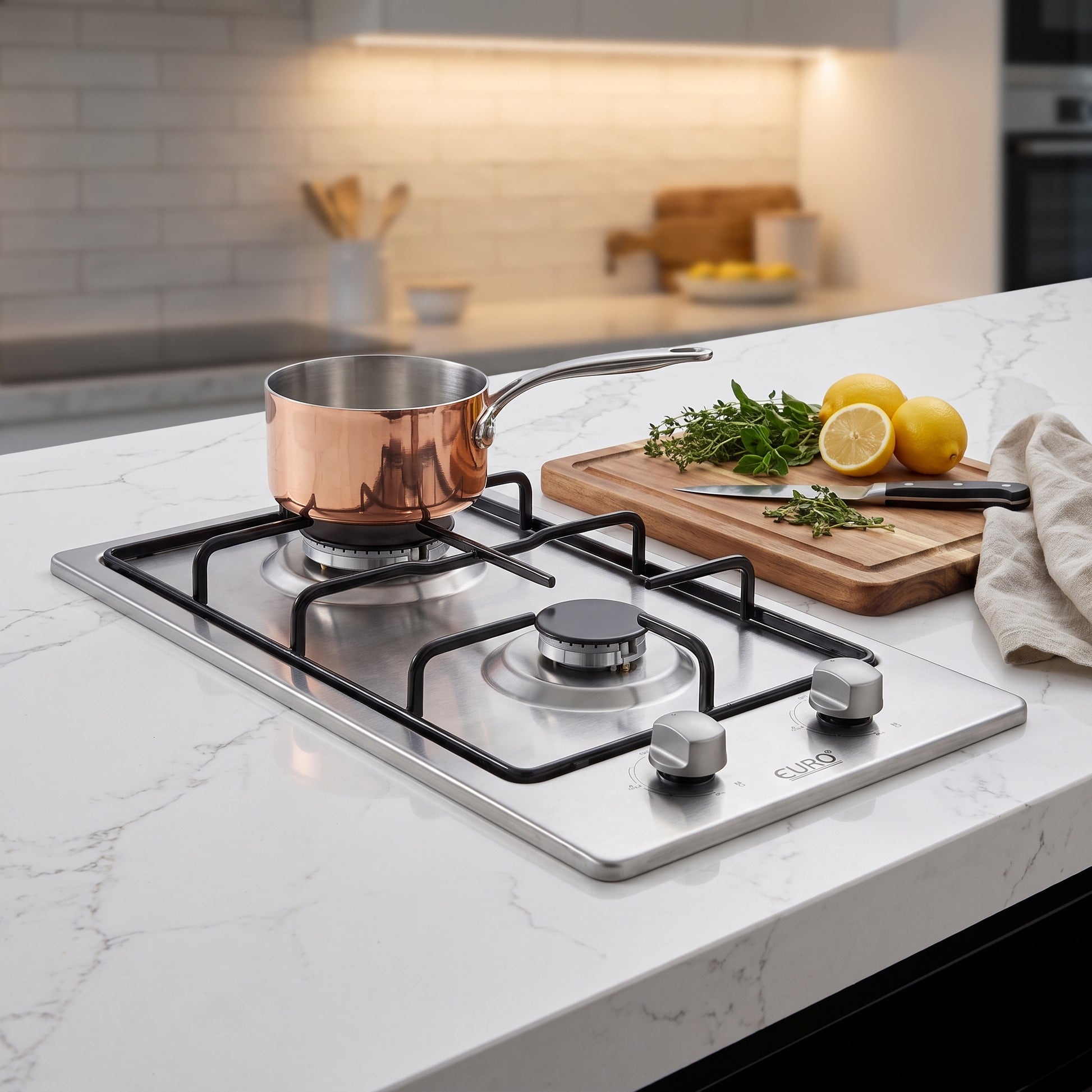 A stainless steel gas cooktop with black grates installed in a white marble kitchen countertop. A copper-colored saucepan sits on one burner, and a wooden cutting board with lemons, herbs, and a knife is placed beside it.
