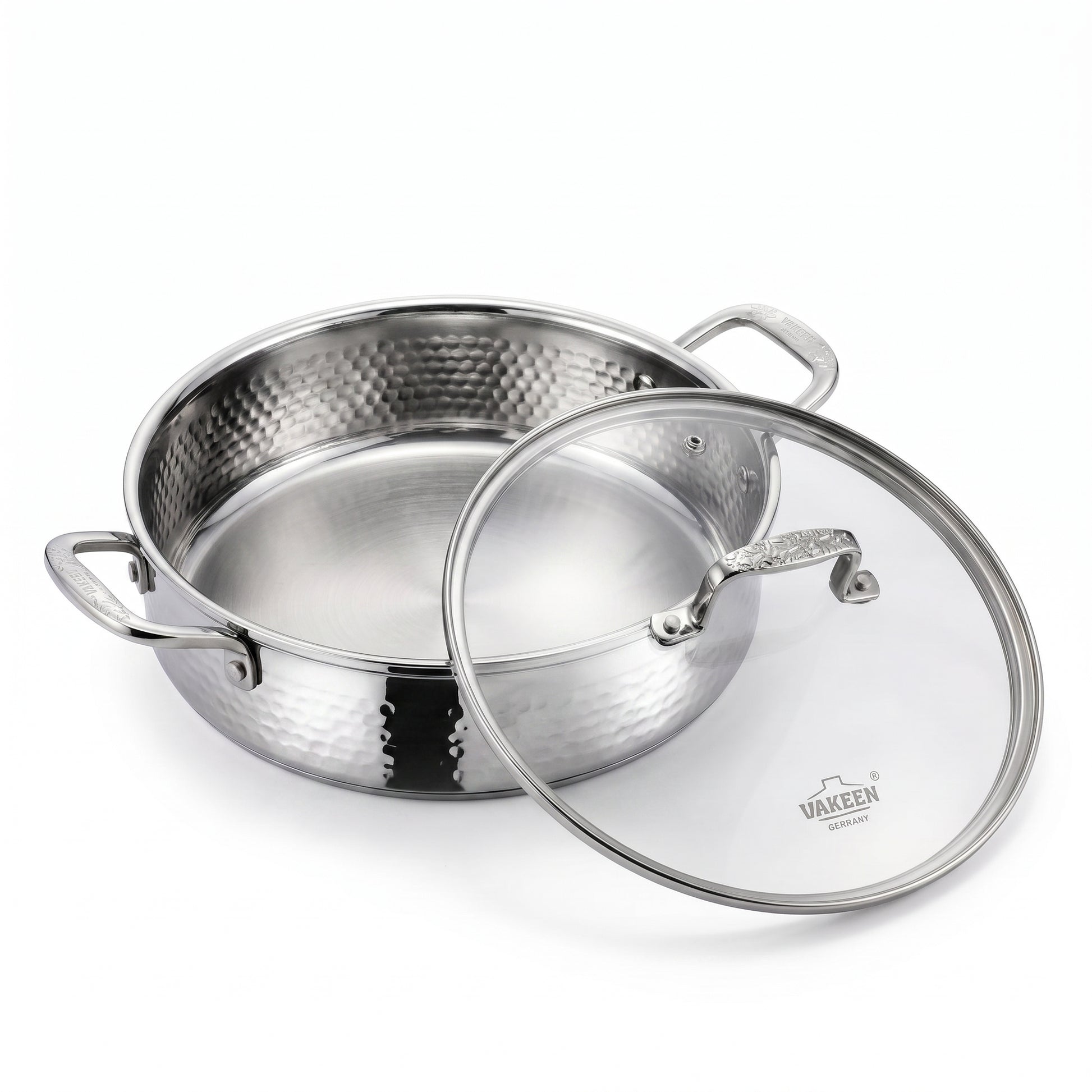 A stainless steel pot with a hammered exterior and a clear glass lid featuring a silver rim and handle, shown on a clean white background. The brand name 'VAKREEN' is visible on the glass lid.