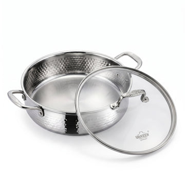 A stainless steel pot with a hammered exterior and a clear glass lid featuring a silver rim and handle, shown on a clean white background. The brand name 'VAKREEN' is visible on the glass lid.