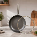 A dark gray nonstick frying pan with a textured interior and a long metallic handle, shown next to a clear glass lid with a metal rim on a kitchen counter.