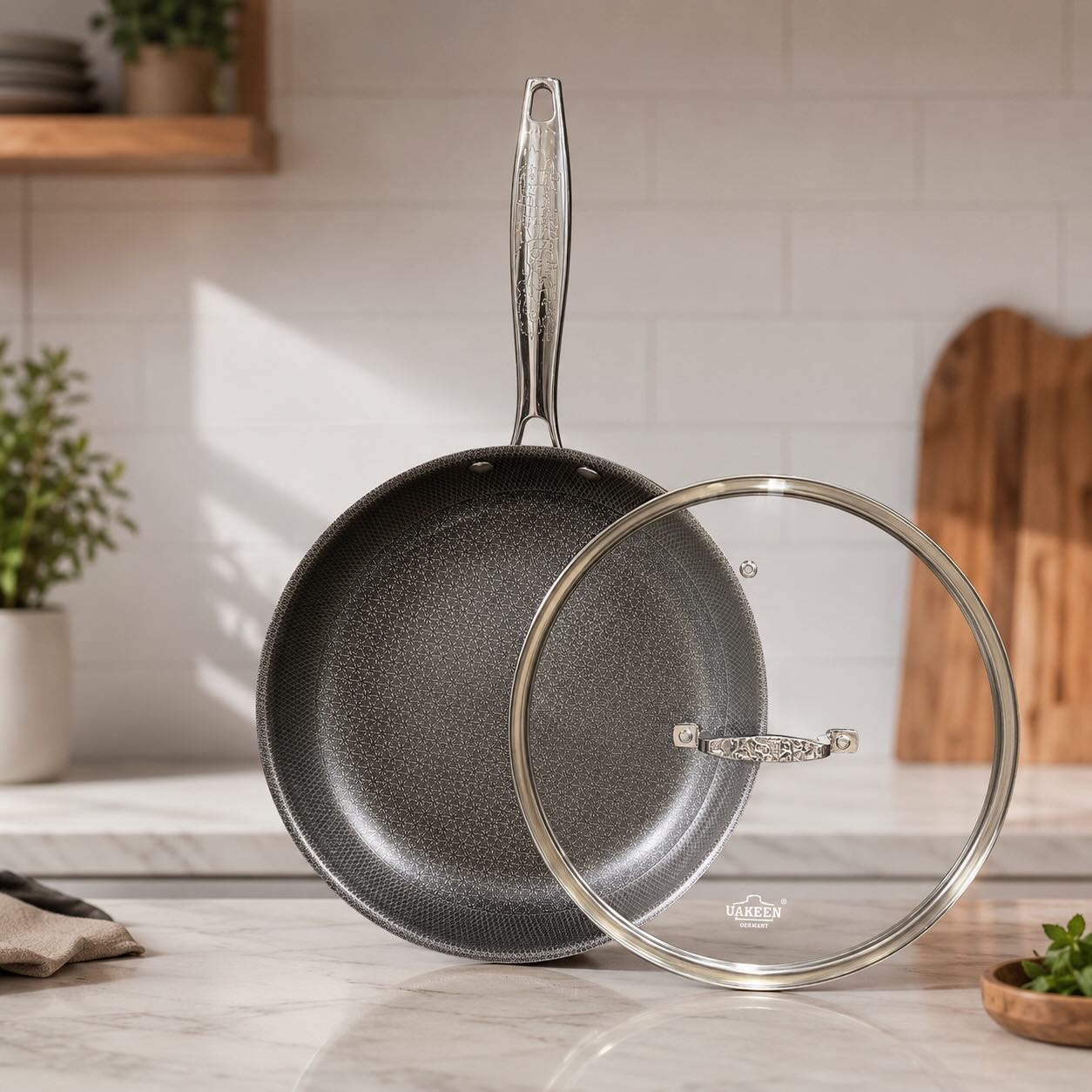 A dark gray nonstick frying pan with a textured interior and a long metallic handle, shown next to a clear glass lid with a metal rim on a kitchen counter.