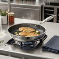 A stainless steel pan with a hammered exterior and a long handle sits on a gas stove burner in a commercial kitchen setting, cooking chicken breasts with garlic and herbs. The background shows stainless steel appliances, spice jars, and a folded dark cloth.